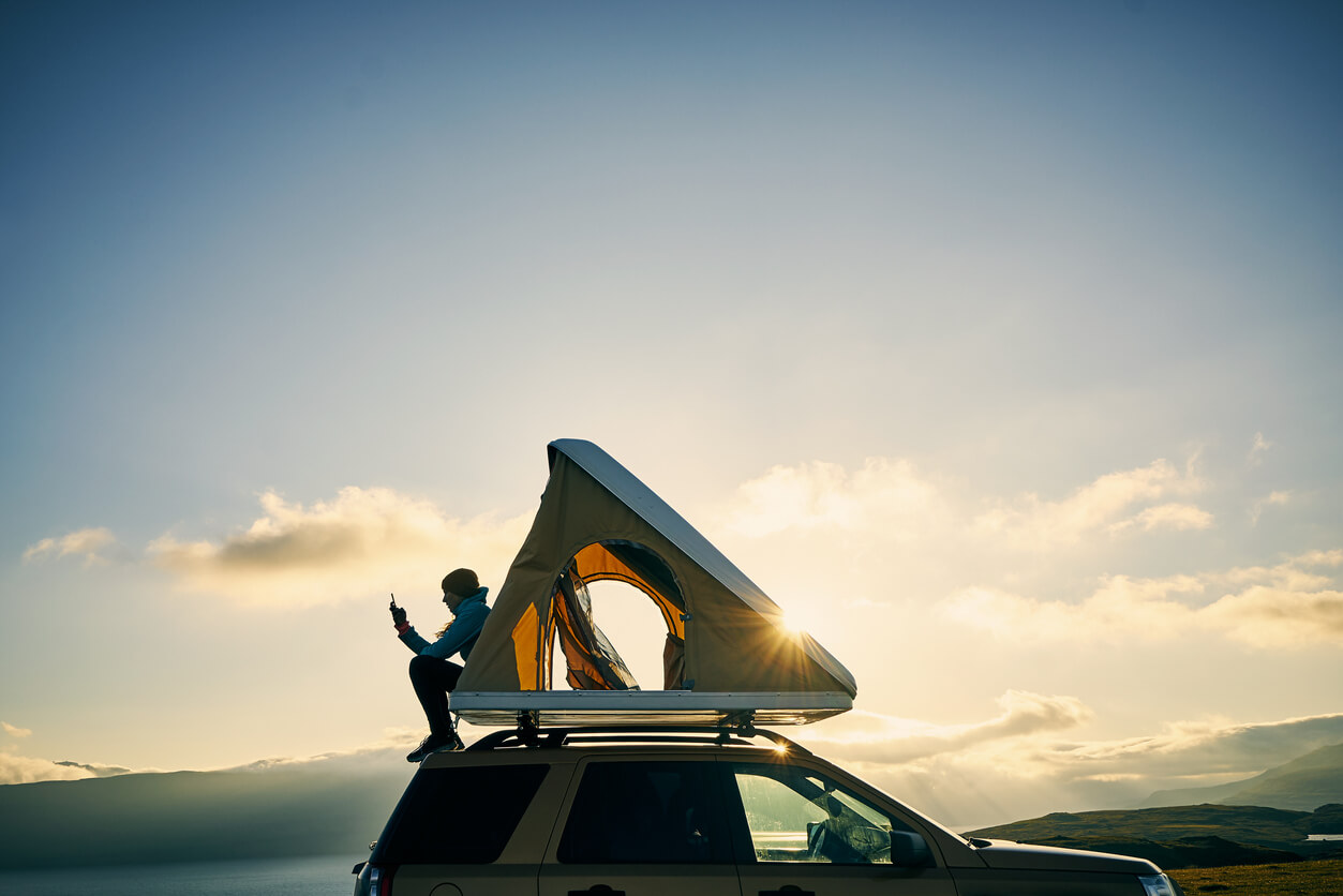 Eine Person sitzt auf einem Autodach mit einem Dachzelt vor einem Sonnenuntergang