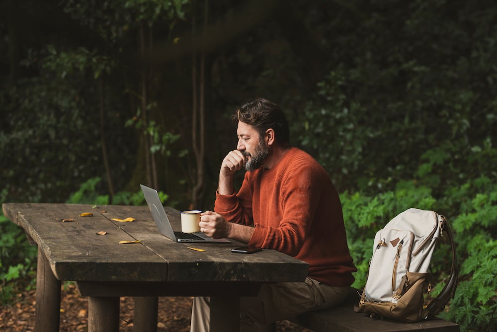 A man sits on a wooden bench at a wooden table in a forrest and looks at his laptop