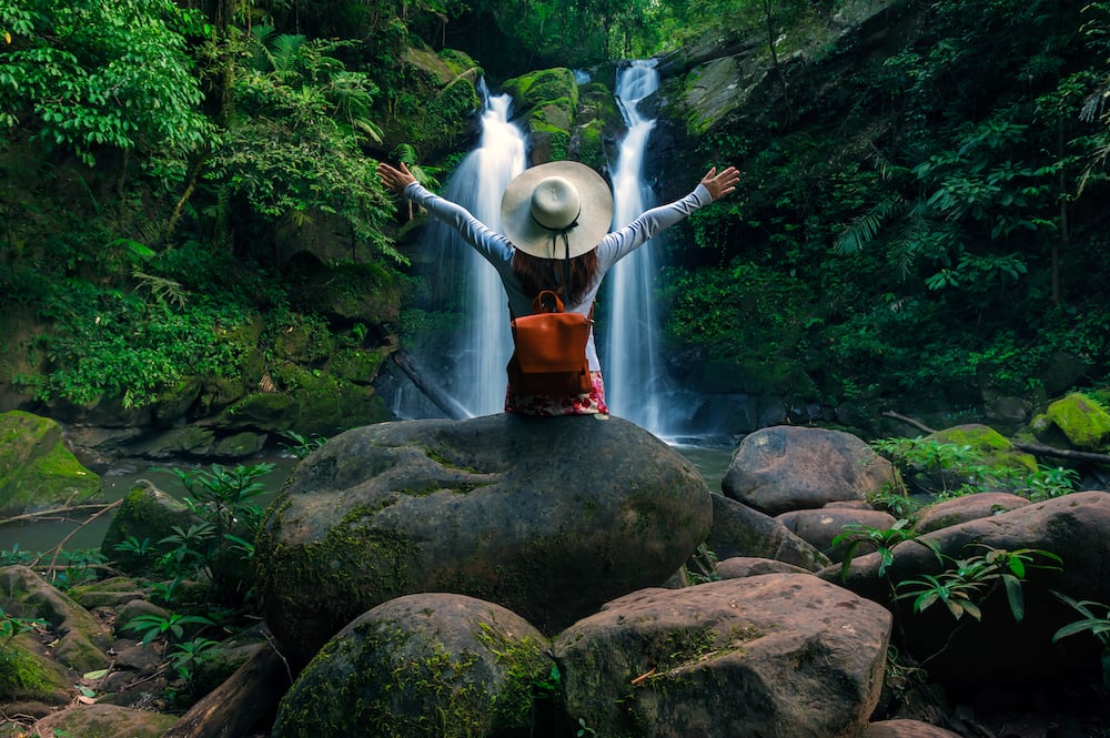 A woman sits on a rock and faces towards a waterfall. She has her arms open in the air. 