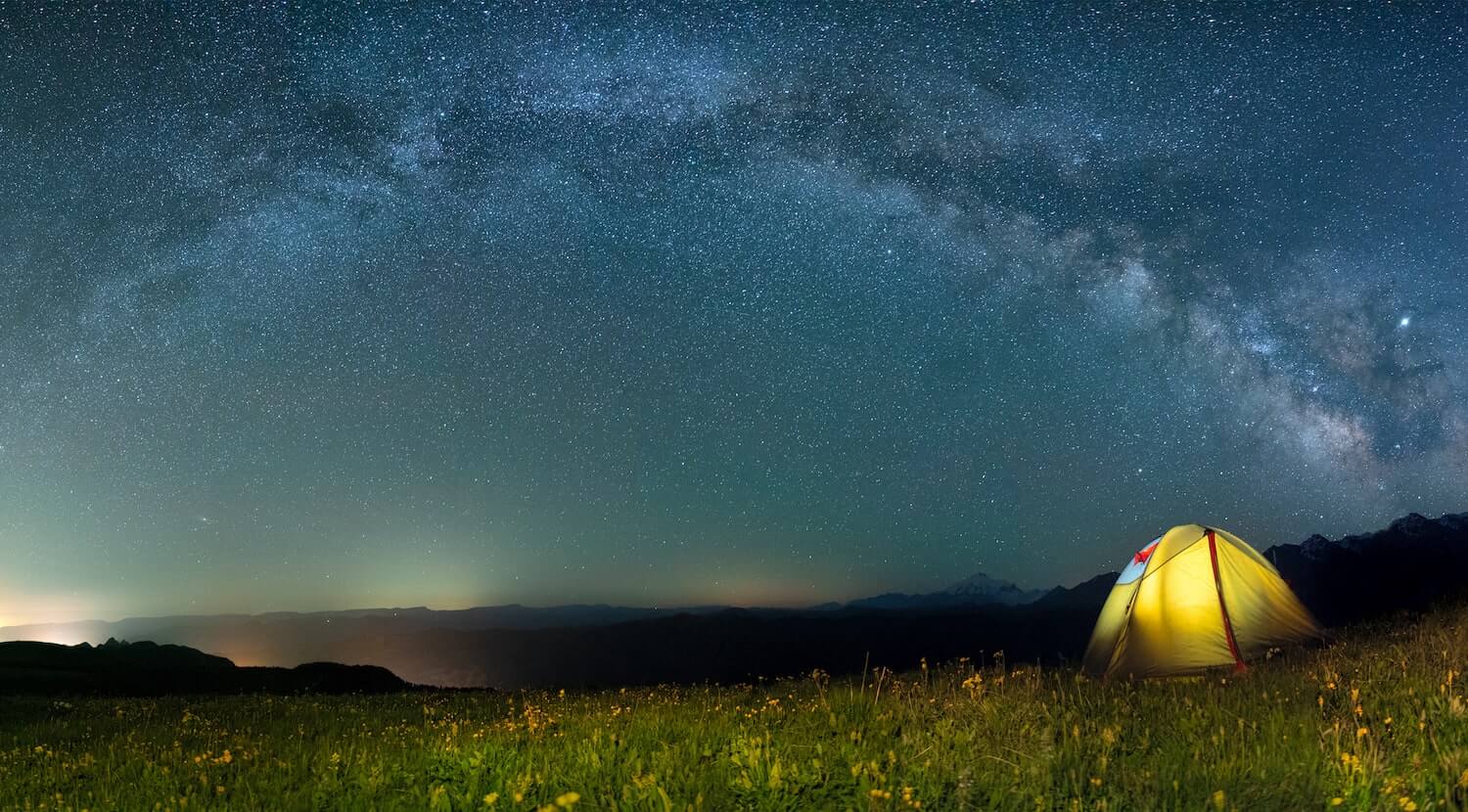A night sky, a flowery field and a tent with a light inside. 